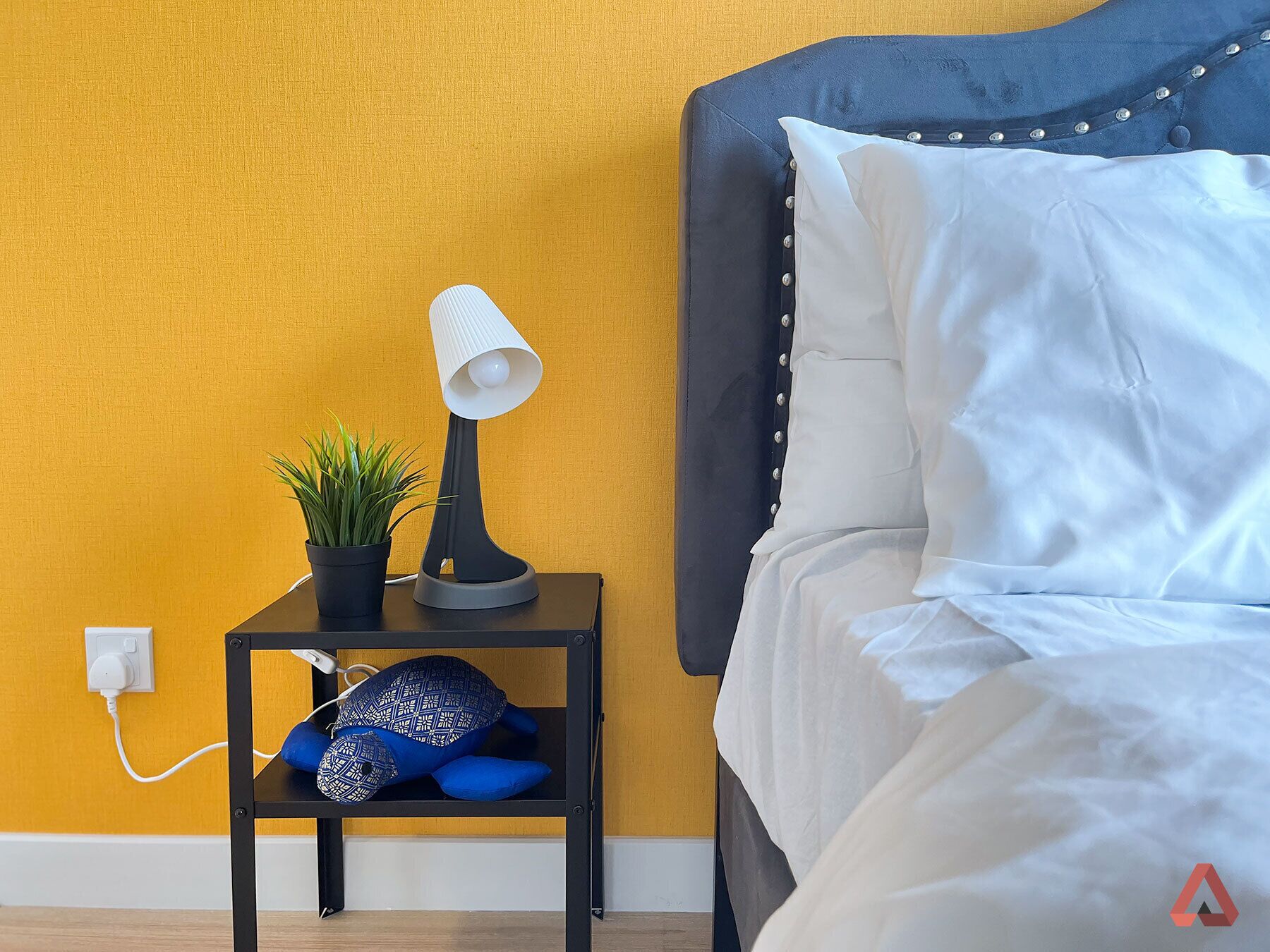 Ein ordentlich bezogenes Bett mit weißen Leinen, eine Nachttischlampe, eine kleine Pflanze und ein blaues Kopfkissen auf einem schwarzen Nachttisch.