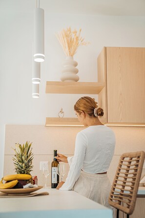 A modern kitchen with a woman pouring wine, a pineapple, bananas, and a bottle of wine on the counter. - Private Villa with Mini Pool, Garden & Parking (Paros)