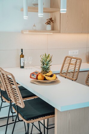 A modern kitchen with a white countertop, wicker chairs, a fruit bowl with pineapple and bananas, wine glasses, and a bottle of wine. - Private Villa with Mini Pool, Garden & Parking (Paros)