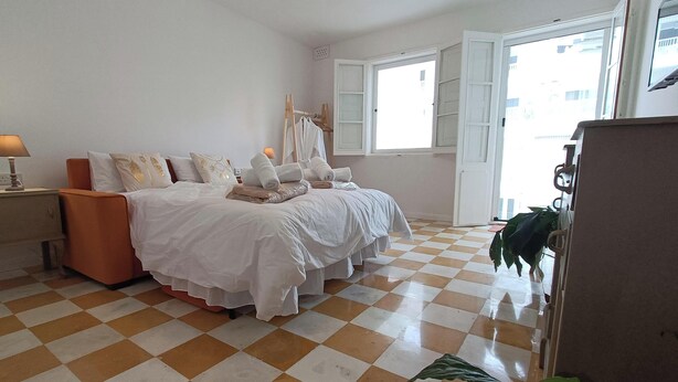 Un dormitorio con piso a cuadros, una cama con ropa de cama blanca, una mesita de noche con lámpara y una ventana con contraventanas abiertas.
