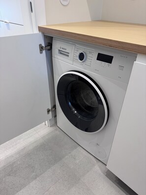 A compact, modern washing machine with a control panel and a wooden countertop above it.