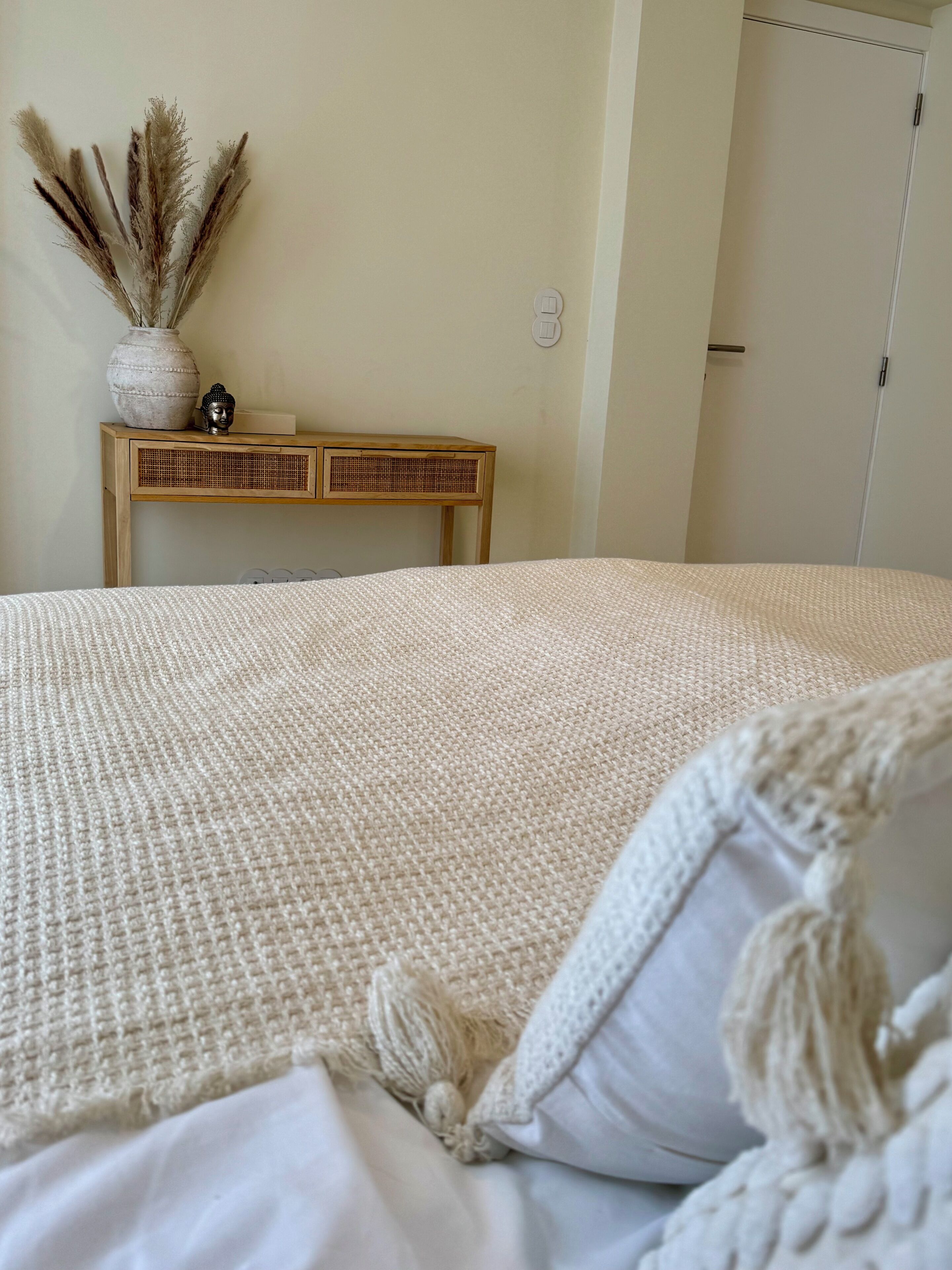 A bed with a textured white cover, a wooden bedside table with a vase and a Buddha statue, and a door in the background.