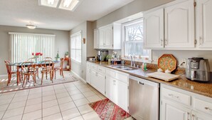 Una cocina con armarios blancos, una mesa de comedor con sillas, una cafetera y un lavavajillas.