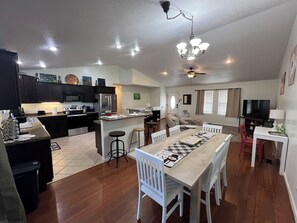 Un vaste espace de vie comprenant une table à manger, des chaises blanches, une cuisine avec des meubles sombres, un réfrigérateur et un ventilateur de plafond.