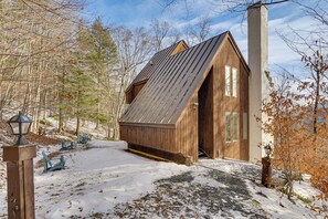A wooden cabin with a sloped roof, a large chimney, and a snow-covered ground.