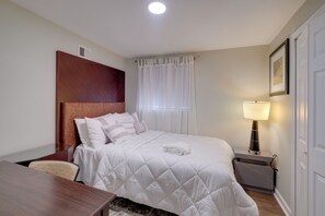 Ein Hotelzimmer mit einem Bett, einem Nachttisch mit Lampe, einem Holz-Kopfteil, einem Fenster mit weißen Vorhängen und einem gerahmten Bild an der Wand.