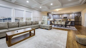 A modern living room with a sectional sofa, a wooden coffee table, and a dining area with chairs and a kitchen in the background. - 1 Mile to Quantico: 5BR Home w/ Game Room! (Triangle)