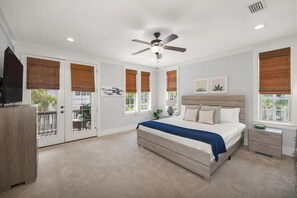 Quarto com cama, mesas de cabeceira, ventoinha no teto e amplas janelas com persianas.