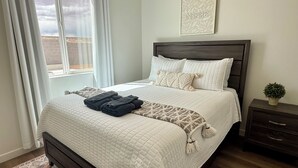 Un dormitorio con cama, mesita de noche, ventana y un cuadro en la pared.