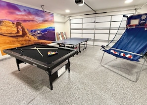 Une table de billard avec des billes, une table de ping-pong et un panier de basketball dans une pièce dont un des murs est décoré d’une fresque représentant un paysage désertique.
