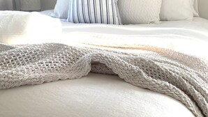 Ein ordentlich bezogenes Bett mit Strickdecke und gestreiften Kissen, einer Nachttischlampe und einer schlichten Wand mit vertikalen Linien.