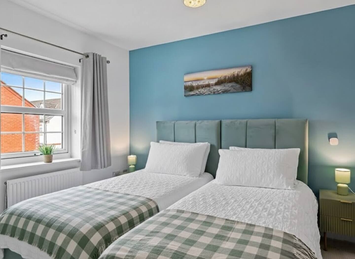 A bedroom with two beds, a window with a view of buildings, a painting on the wall, and bedside lamps.