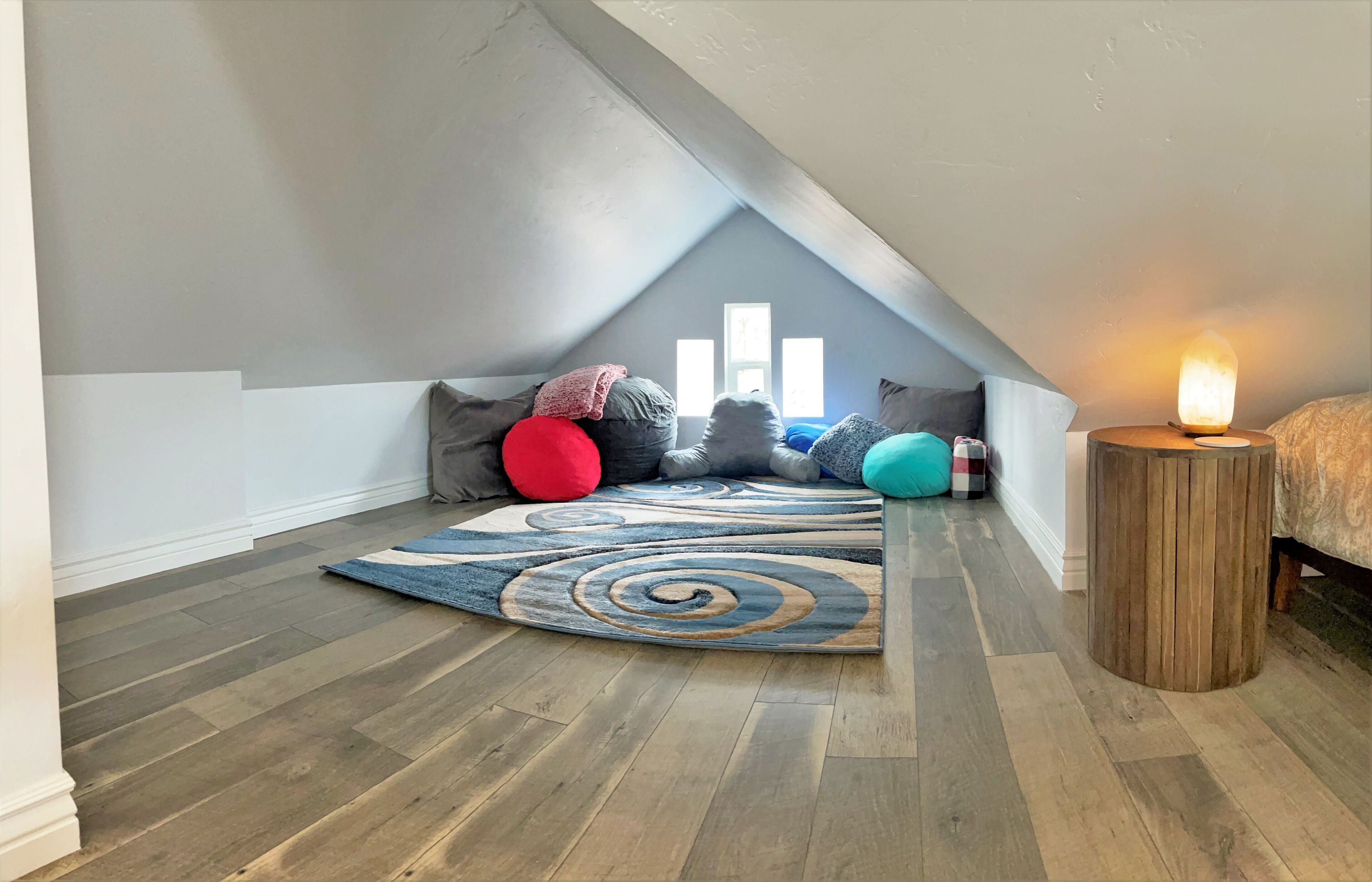 A cozy attic bedroom with a sloped ceiling, a wooden floor, a colorful spiral rug, and a small table with a candle.