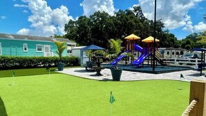 Ein Spielplatz mit Rutschen und einer Spielkonstruktion, umgeben von einer gepflegten Rasenfläche und einem Haus im Hintergrund.
