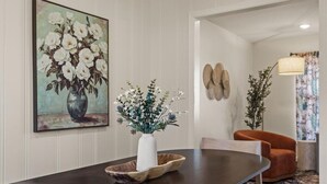 Un comedor con una mesa de madera oscura, un jarrón blanco con flores y un cuadro de flores blancas en la pared.