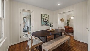 Un comedor con una mesa de madera oscura, sillas blancas y un banco. Hay un cuadro en la pared y una planta en el suelo.