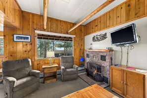 A cozy cabin interior with wooden walls, a fireplace, a TV mounted on the wall, and two armchairs. - Glorious, dog friendly, 2 bedroom cabin with great views close to RMNP! (Estes Park)