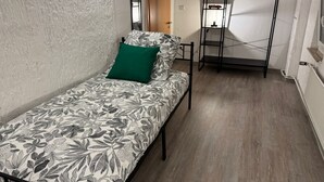 Quarto pequeno com cama de solteiro, colcha com padrão floral, almofada verde e escada de metal preta encostada à parede.