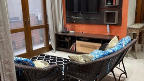 Una sala de estar con un televisor de pantalla plana grande montado en la pared, un sofá de mimbre con cojines estampados, una pequeña mesa de madera y una estantería decorativa con varios objetos.