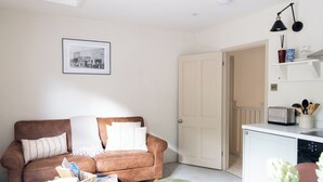 A living room with a brown sofa, a small table, a framed picture on the wall, a door, and a kitchenette area. - Garden apartment with balcony (Bath)