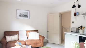 A living room with a brown sofa, a small table, a framed picture on the wall, a door, and a kitchenette area.