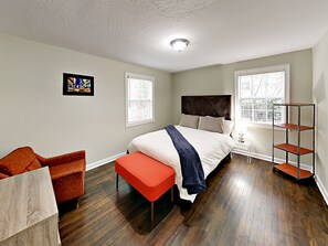 Ein Schlafzimmer mit einem Bett, einem Sessel, einer Bank, einem Regal und zwei Fenstern.