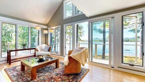 A spacious living room with large windows, a wooden coffee table, a sofa, and a chair. - Kezar Lakefront Home: 2 Boat Dock & Hot Tub (Lovell)
