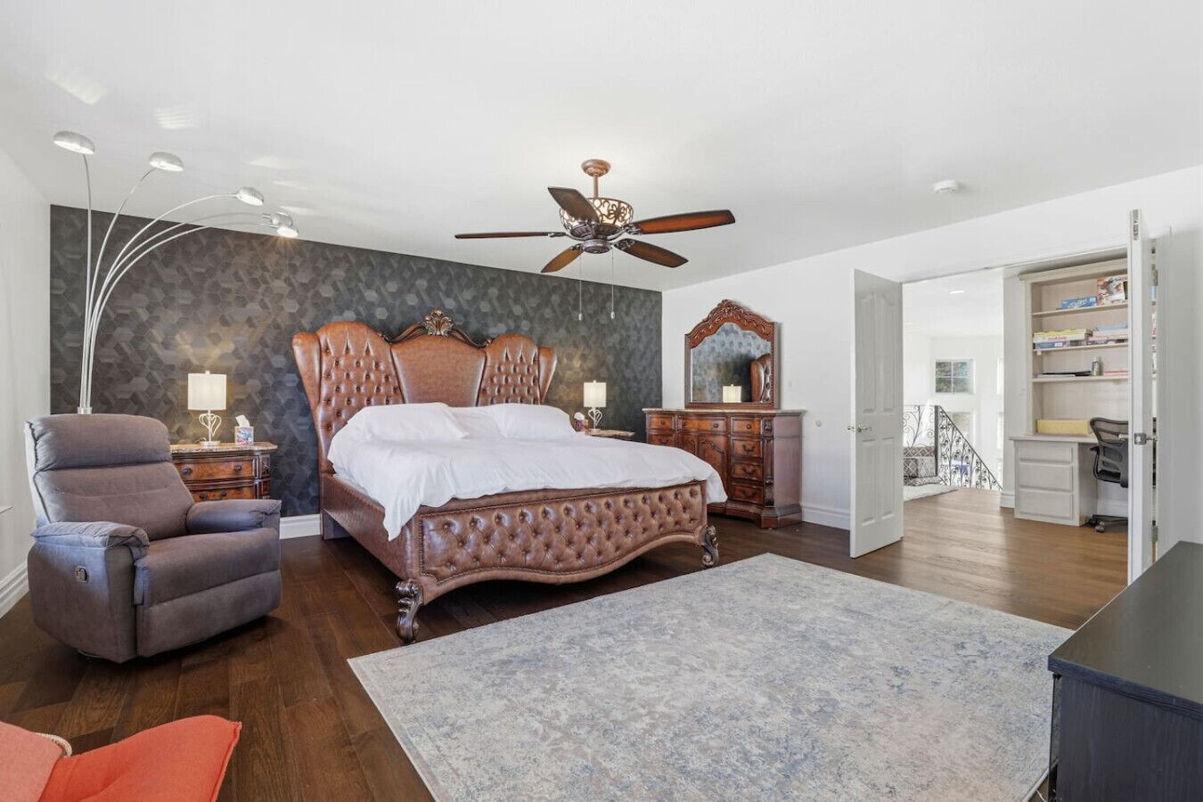 A bedroom with a large bed, a ceiling fan, a nightstand, a dresser, and a desk with a chair.