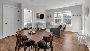 Een ruime woonkamer met een eettafel, stoelen, een grijze bank en een aan de muur bevestigde televisie.