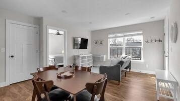 A spacious living area with a dining table, chairs, a gray sofa, and a TV mounted on the wall.