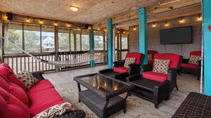 A covered outdoor seating area with wicker furniture, red cushions, and a hammock. - Stunning OBX 6BR Estate, Pool, Spa, Game Room, Fire Pit & Volleyball (Salvo)