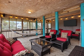 A covered outdoor seating area with wicker furniture, red cushions, and a hammock. - Stunning OBX 6BR Estate, Pool, Spa, Game Room, Fire Pit & Volleyball (Salvo)