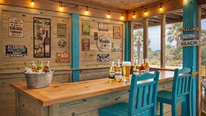 A wooden bar area with a wooden counter, blue chairs, and a bucket of beer bottles. - Stunning OBX 6BR Estate, Pool, Spa, Game Room, Fire Pit & Volleyball (Salvo)
