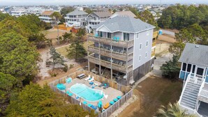A multi-story residential building with a backyard swimming pool, surrounded by trees and other houses. - Stunning OBX 6BR Estate, Pool, Spa, Game Room, Fire Pit & Volleyball (Salvo)