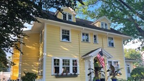 A yellow house with a white picket fence, a front porch with potted plants, and an American flag. - Elegant Historic Home near University of Rochester (Rochester)