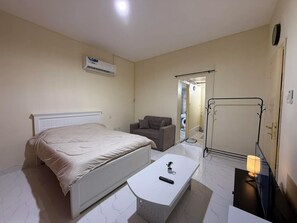 A bedroom with a bed, a chair, a small table with a remote control, a lamp, and a wardrobe. - Perfect 1-bedroom villa with AC in charming Abu Dhabi (Abu Dhabi)