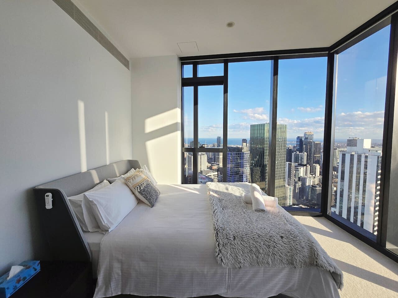 A modern bedroom with a large bed, a nightstand, and a view of a cityscape through large windows.