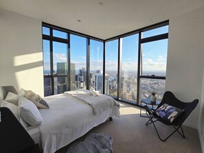 Ein modernes Schlafzimmer mit einem großen Bett, einem Sessel und Blick auf eine Stadtlandschaft durch raumhohe Fenster.