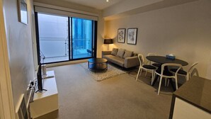 Ein modernes Wohnzimmer mit einer Couch, einem runden Couchtisch, einem Esstisch mit Stühlen, einem Fernseher und einem großen Fenster mit Blick auf Gebäude.