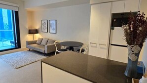 Eine moderne Küche mit weißen Schränken, einer schwarzen Arbeitsplatte und einer Vase mit Trockenblumen. Es gibt einen Essbereich mit Tisch und Stühlen, und eine Couch im Hintergrund.