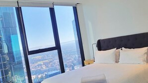 Une chambre d’hôtel avec un grand lit, une tête de lit sombre et une vue sur le paysage urbain grâce à de grandes fenêtres du sol au plafond.