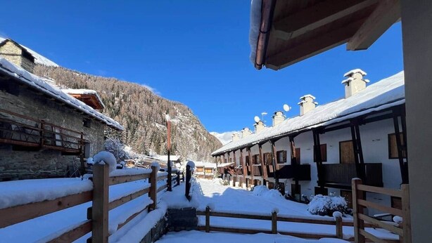 Un sentier enneigé mène à une rangée de bâtiments traditionnels en bois, dont les toits sont recouverts de neige, sous un ciel bleu dégagé.