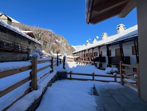 Ein verschneiter Pfad führt zu einer Reihe traditioneller Holzhäuser mit Schnee auf den Dächern und blauem, klarem Himmel.