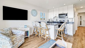 Een moderne keuken met witte kasten, een magnetron en een fornuis. Er staan rieten stoelen en een klein eiland met krukken. Een flatscreen televisie hangt aan de muur en er is een klok te zien.