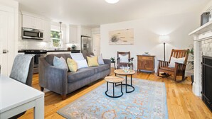 Eine moderne Küche mit weißen Schränken, einem grauen Sofa mit Kissen, einem Holzschaukelstuhl und einem gemusterten Teppich.