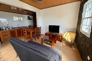A compact living space with a sofa, TV, dining table, chairs, kitchen area, and a large window. - Goat's Retreat (São Mateus)