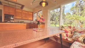 A kitchen with wooden cabinets, a granite countertop, and a large window with a view of trees. - Serene country home in charming Volcano Village near the Volcano National Park. (Volcano)