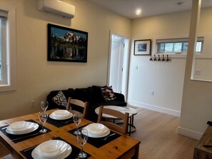 A dining area with a wooden table set for four, a black sofa with a patterned pillow, and a framed picture of a mountain landscape on the wall. - New 2BR Garden Suite with AC and private patio (Burnaby)