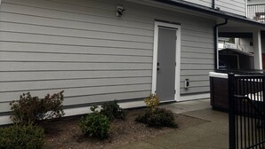 A two-story building with a grey siding exterior, a front door, and a small porch area. - New 2BR Garden Suite with AC and private patio (Burnaby)