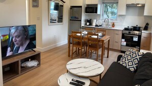 A modern living room with a TV, a dining area with chairs and a table, a kitchen with appliances, and a sofa with a patterned pillow. - New 2BR Garden Suite with AC and private patio (Burnaby)